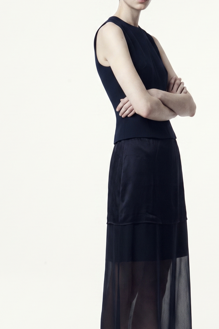 A model wearing a sleeveless navy dress with a structured bodice and a layered, semi-sheer skirt, shown from the side against a plain white studio background. view 1