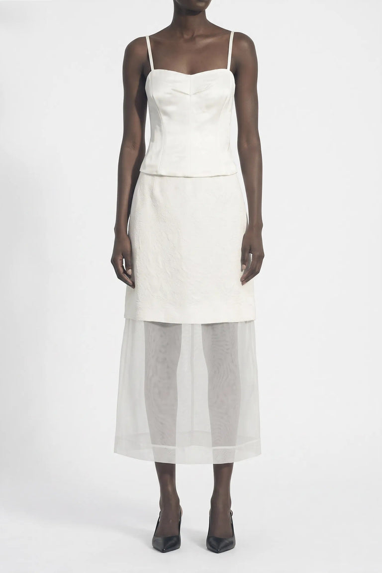 A model wearing a white sleeveless midi dress with a structured corset-style bodice, a defined waist seam, and a sheer white tulle overskirt, paired with black pointed-toe heels against a plain white studio background. view 2